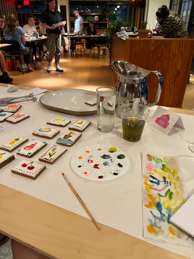 table with water-color painting supplies: a brush, paint palate, and several tiny paintings except the paintings are also cookies